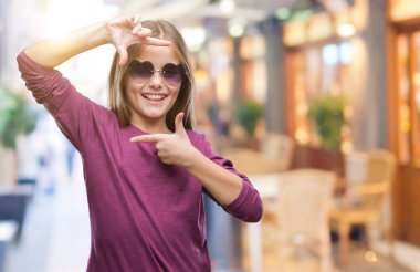 Genç güzel kız elle yapma çerçeve ve parmakları ile mutlu yüz gülümseyen izole arka plan üzerinde güneş gözlüğü takıyor. Yaratıcılık ve Fotoğrafçılık kavramı.