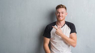 Genç Kızıl saçlı adam gri grunge duvarın neşeli bir gülümseme ile el ve parmak kadar kameraya bakarak yüzündeki mutlu ve doğal ifade ile yan ile işaret yüz üzerinden.