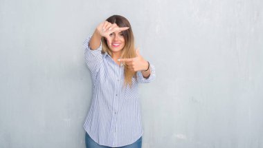 Gri grunge duvar elle yapma çerçeve ve parmakları ile mutlu yüz gülümseyen moda iş kıyafeti giymiş genç yetişkin kadın. Yaratıcılık ve Fotoğrafçılık kavramı.
