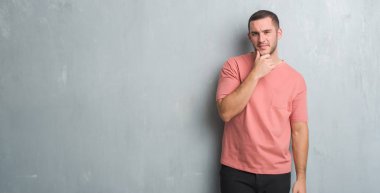 Beyaz delikanlı emin çapraz kollar ve çeneme kaldırdı el ile gülümseme ile kamera seyir gri grunge duvarın üzerinden. Pozitif düşünce.