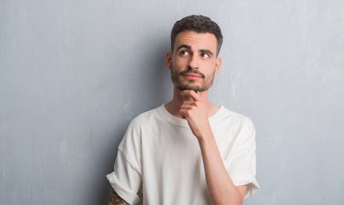 Genç yetişkin adam gri grunge duvar ciddi bir yüz düşünme hakkında soru, çok karışık bir fikir üzerinde ayakta