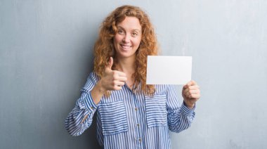 Gri grunge genç Kızıl saçlı kadın boş kartı ok işareti yapıyor büyük gülümseme ile mutlu tutarak duvar, parmaklarıyla, mükemmel işaret parmak