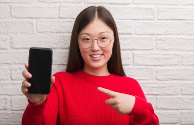 Tuğla duvar gösteren smartphone el ve parmak ile işaret çok mutlu genç Çinli kadın