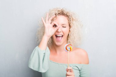 Sarışın genç kadın ile mutlu yüz el parmaklarının arasından seyir göz üzerinde ok işaretiyle yapıyor gülümseyen candy lolipop yemek grunge gri duvarın üzerinden