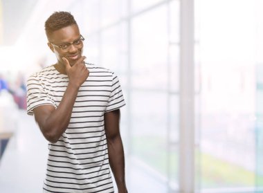Gözlük ve emin çapraz kollar ve çeneme kaldırdı el ile gülümseme ile kameraya bakarak lacivert t-shirt giyen genç Afro-Amerikan adam. Pozitif düşünce.