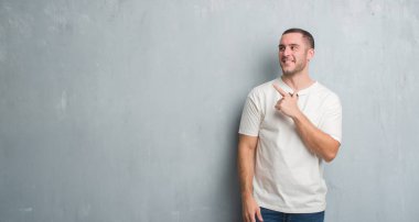 Genç beyaz adam neşeli bir gülümseme ile el ve parmak kadar mutlu ve doğal ifade kameraya bakarak yüzünde yanında işaret yüz ile güneş gözlüğü takıyor gri grunge duvarın üzerinden.