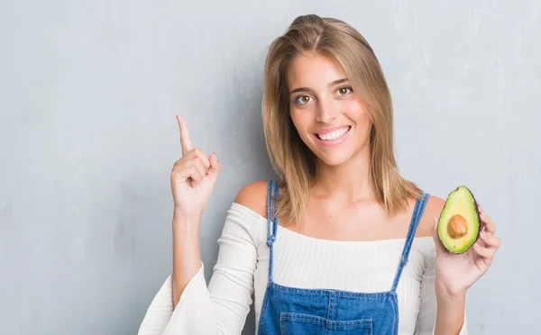 Hermosa Mujer Joven Sobre Pared Gris Grunge Comiendo Aguacate Sorprendido —  Fotos de Stock