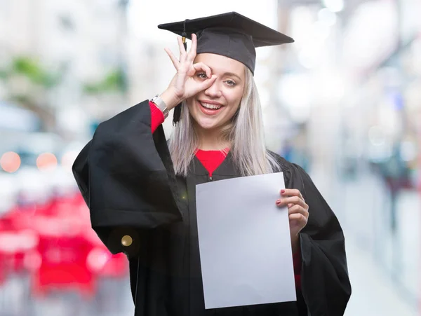 指の間から見て目に手で サインをして笑っている幸せそうな顔で孤立の背景の上の学位を保持する大学院の制服を着て若いブロンドの女性 — ストック写真