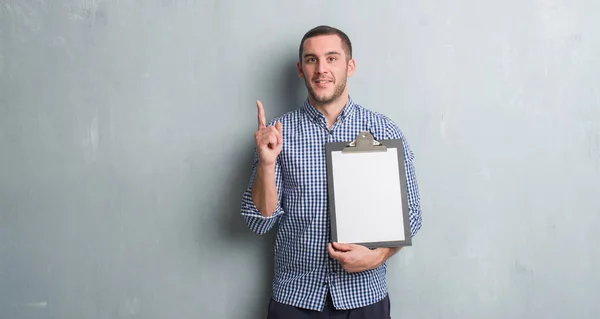 Young Caucasian Man Grey Grunge Wall Holding Clipboard Surprised Idea — Stock Photo, Image