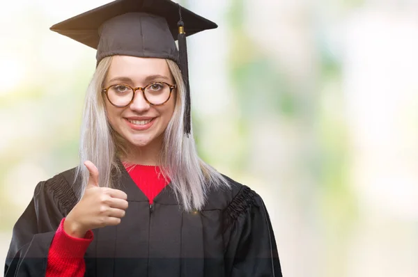 若いブロンドの女性は 幸せな親指の手でジェスチャーを行う分離の背景に大学院の制服を着ています 成功を示すと カメラを見て式を承認します — ストック写真