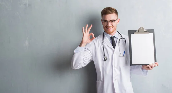 Jovem Médico Ruivo Homem Sobre Parede Grunge Cinza Segurando Prancheta — Fotografia de Stock