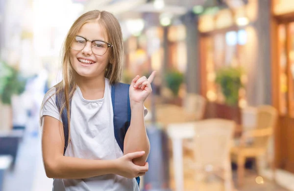 若く美しくスマートな学生の女の子の顔に大きな笑みを浮かべて隔離された背景にバックパックを着てカメラを見て側の手と指で指す — ストック写真