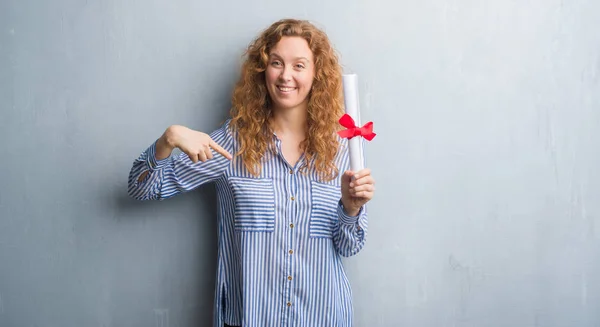 Young redhead business woman over grey grunge wall holding diploma with surprise face pointing finger to himself