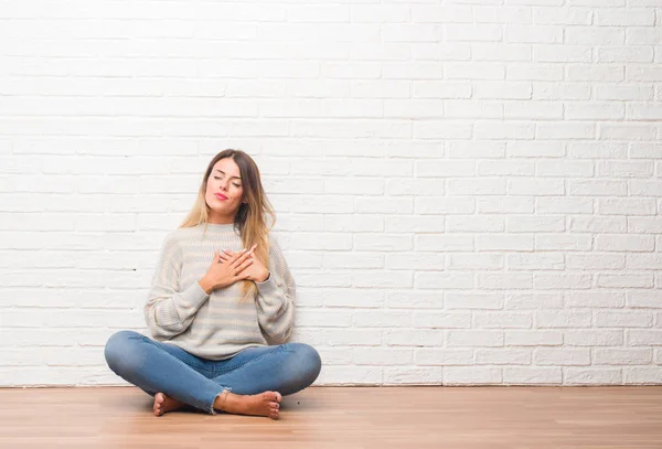 自宅の白いレンガ壁に床に座って若い成人女性を浮かべて目を閉じて胸に手と顔に感謝のジェスチャー 健康の概念 — ストック写真
