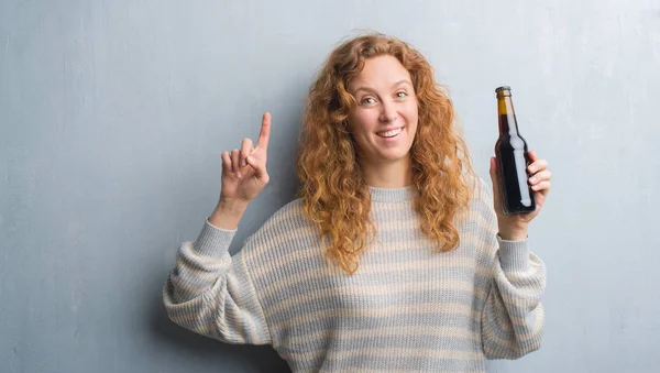 Молодая Рыжая Женщина Над Серой Гранж Стеной Держа Бутылку Пива — стоковое фото