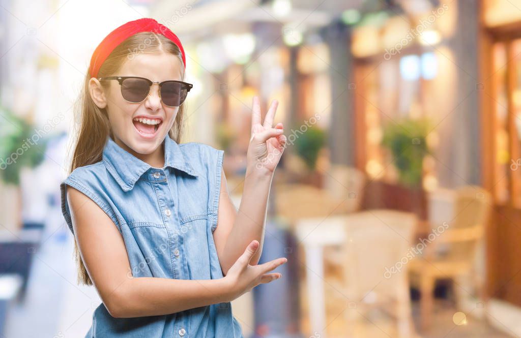 Young beautiful girl wearing sunglasses over isolated background smiling with happy face winking at the camera doing victory sign. Number two.