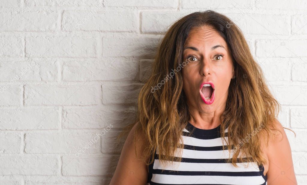 Middle age hispanic woman over white brick wall scared in shock with a surprise face, afraid and excited with fear expression