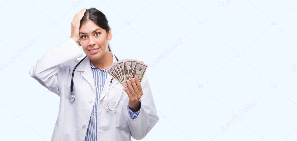 Young hispanic doctor woman holding dollars stressed with hand on head, shocked with shame and surprise face, angry and frustrated. Fear and upset for mistake.