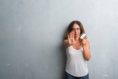 Açık eliyle dur işareti ile ciddi ve kendine güvenen ifade, savunma jest yapıyor gri grunge duvar holding kredi kartı üzerinde duran orta yaş İspanyol kadın