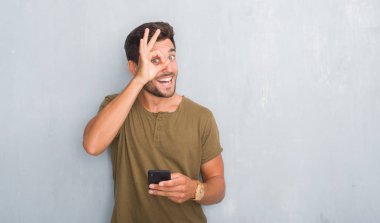 Yakışıklı genç gri grunge üzerinde duvar manifatura mutlu yüz el parmaklarının arasından seyir göz üzerinde ok işaretiyle yapıyor gülümseyen ile smartphone kullanarak bir ileti