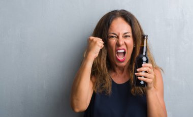 Orta yaş İspanyol kadın bira şişesi holding gri grunge duvardan ayakta rahatsız ve boşa çıkarmak ile öfke, çılgın da yükseltilmiş el ile bağırmaya kavramı öfke