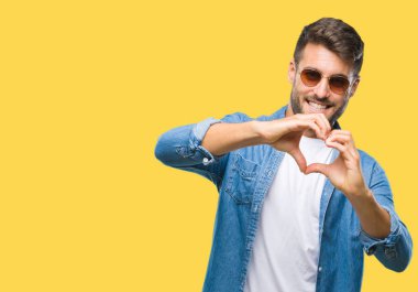 Genç yakışıklı adam aşık kalp simgesi ve elleriyle şekil gösteren gülümseyen izole arka plan üzerinde güneş gözlüğü takıyor. Romantik kavramı.