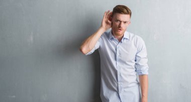 Genç redhead iş adamı bir işitme söylenti ya da dedikodu için dinleme kulak üzerinde el ile gülümseyen gri grunge duvarın üzerinden. Sağırlık kavramı.