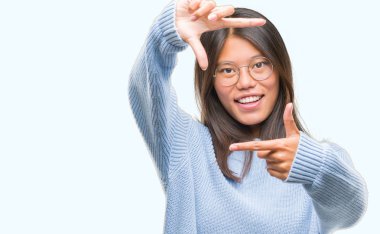 Genç Asyalı kadın elle yapma çerçeve ve parmakları ile mutlu yüz gülümseyen izole arka plan üzerinde kış kazak giyiyor. Yaratıcılık ve Fotoğrafçılık kavramı.