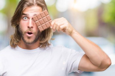 Uzun saç beslenme ile yakışıklı delikanlı çikolata bar izole arka şok sürpriz yüzü, korkuyor ve korku ifadesi ile heyecanlı olan korkmuş üzerinden