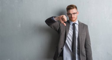 Genç redhead zarif iş adamı kızgın yüz, gri grunge duvarın üzerinden negatif antipati başparmak aşağı, ret kavramı ile gösterilen işareti