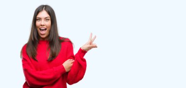 Genç güzel İspanyol mutlu yüz zafer işareti yaparken kameraya göz kırpıyor ile gülümseyen kırmızı kazak giyiyor. İki numaralı.