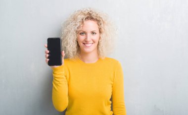 Grunge gri arka plan ayakta ve diş gösterilen kendine güvenen bir gülümsemeyle gülümseyerek mutlu bir yüz ile akıllı telefon boş ekran gösteren genç sarışın kadın