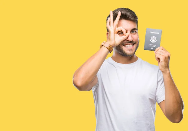 年轻英俊的人持有美国护照在孤立的背景与快乐的脸微笑做 签名用手在眼睛看通过手指 — 图库照片