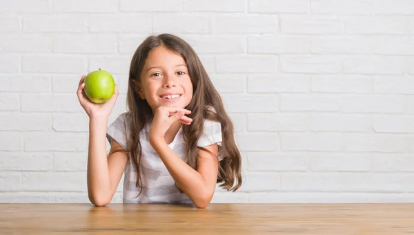 立っていると自信を持って歯を見せて笑顔で笑っている幸せそうな顔で新鮮な青リンゴを食べるテーブルに座って若いヒスパニックの子供 — ストック写真