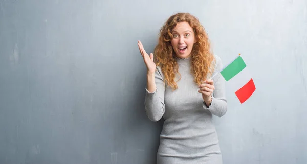 灰色グランジ壁保有上の若い赤毛の女性イタリア非常に幸せと興奮 笑顔で叫び 手を上げた勝者式祝う勝利のフラグします — ストック写真