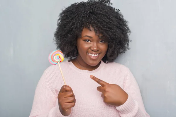 Young african american woman over grey grunge wall eating lollipop candy very happy pointing with hand and finger