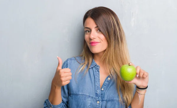灰色垃圾墙上的年轻成年妇女吃新鲜的绿色苹果愉快与大微笑做 拇指用手指 优秀标志 — 图库照片