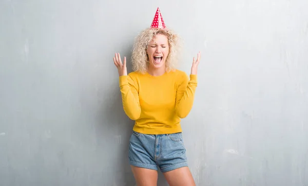 グランジに若いブロンドの女性灰色の狂牛病を祝う誕生日帽子を身に着けている壁と腕の成功のために狂気悲鳴上げられたとクローズの目興奮します 勝者の概念 — ストック写真