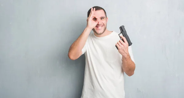 年轻的白种人犯罪男子在灰色的垃圾墙持有枪与快乐的脸微笑做 标志用手在眼睛看通过手指 — 图库照片