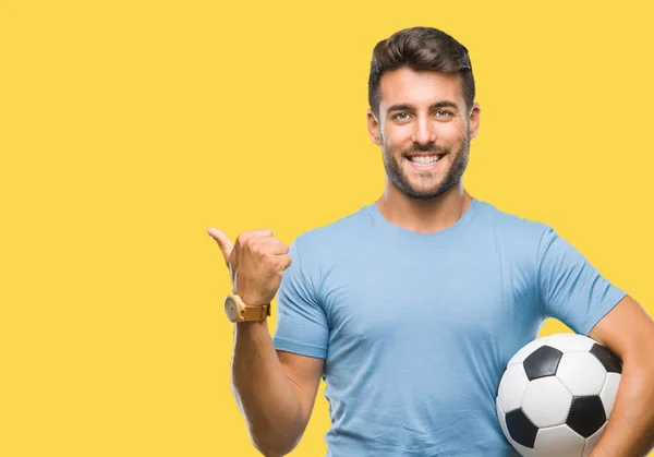 年轻英俊的男子拿着足球足球在孤立的背景指向和显示与拇指到一边 快乐的脸微笑 — 图库照片