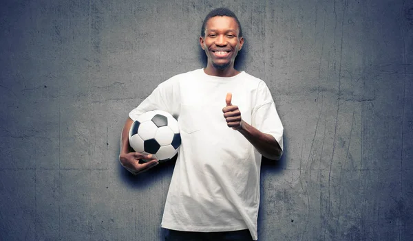 広く示す笑みを浮かべてサッカー ボールを保持しているアフリカの黒人男性の親指カメラのような式および承認するジェスチャを — ストック写真