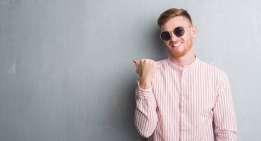 Genç Kızıl saçlı adam işaret ve başparmak kadar mutlu yüz gülümseyen tarafı ile gösterilen retro güneş gözlüğü takıyor gri grunge duvarın üzerinden