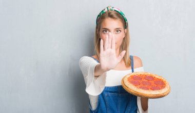 Grunge gri duvar açık eliyle dur işareti ile ciddi ve kendine güvenen ifade, savunma jest yapıyor pepperoni pizza yemek güzel genç kadın