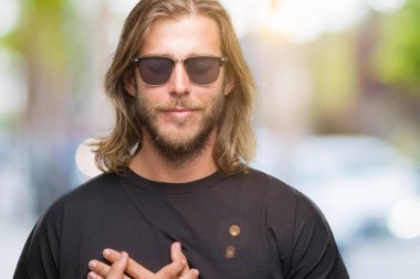 Göğüs kapalı gözler ve yüzünde minnettar jest ile gülümseyen izole arka plan üzerinde güneş gözlüğü takmış uzun saçlı yakışıklı delikanlı eller. Sağlık kavramı.