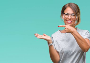 Hayret ve el ile sunulması ve parmak ile işaret ederken kameraya gülümseyen izole arka plan üzerinde gözlük takan orta yaş İspanyol kadın kıdemli.