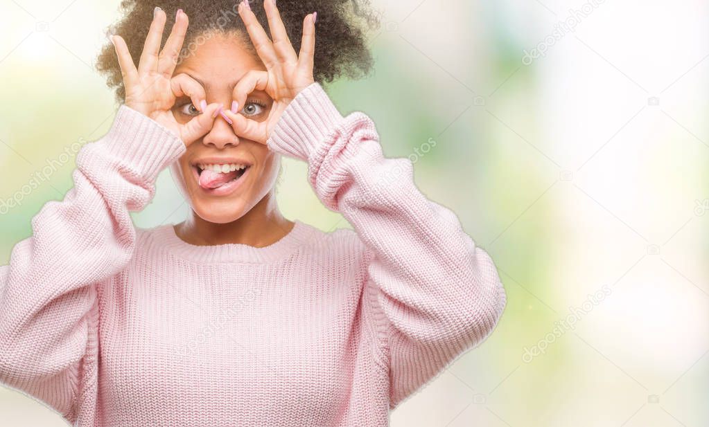 Young afro american woman wearing winter sweater over isolated background doing ok gesture like binoculars sticking tongue out, eyes looking through fingers. Crazy expression.