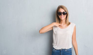 Sürpriz yüz işaret parmağı kendine moda güneş gözlüğü takıyor grunge gri duvar üzerinde duran güzel genç kadın