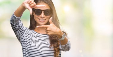 Genç güzel Arap kadın elle yapma çerçeve ve parmakları ile mutlu yüz gülümseyen izole arka plan üzerinde güneş gözlüğü takıyor. Yaratıcılık ve Fotoğrafçılık kavramı.