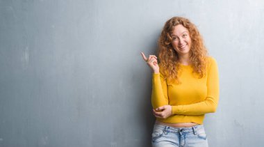 Yüzünde kocaman bir gülümseme ile gri grunge duvarın üzerinden kameraya bakarak yan el ve parmak ile işaret eden genç Kızıl saçlı kadın.