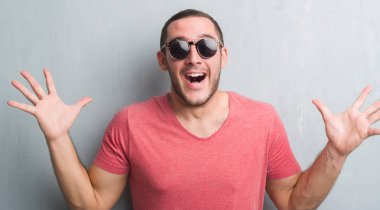 Gri grunge üzerinde beyaz delikanlı duvar büyük gülümseme ile çığlık zaferini kutluyor ve ellerini kaldırdı giyen güneş gözlüğü çok mutlu ve heyecanlı, kazanan ifade